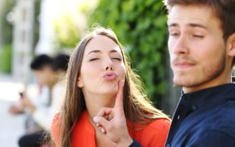 A woman with long hair puckers her lips as if to kiss, while a man lightly touches her chin with his finger, looking away with a smirk. They appear to be outdoors, with greenery in the background and two blurred figures in the distance.