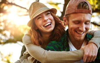 A woman with red hair, wearing a hat, joyfully piggybacks on a smiling man in a cap. Both appear happy and relaxed, enjoying the outdoors with sunlight filtering through trees in the background.