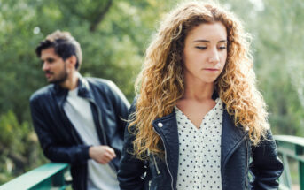 A woman with curly blonde hair and wearing a polka dot blouse and black leather jacket stands in the foreground, looking to the side. In the background, a man with short dark hair and a beard, also in a leather jacket, leans on a green railing, looking away.