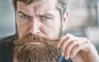 A bearded man with blue eyes intensely gazes into the camera while twirling a strand of his mustache with his right hand. The background is blurred, focusing the attention on his expressive face.