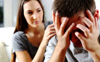 A woman with long brown hair and wearing a gray shirt gently places her hand on the shoulder of a man who is holding his face in his hands, showing he is distressed. She looks concerned while trying to comfort him.