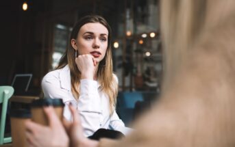 A young woman with long, blonde hair, wearing a white shirt, sits in a cafe with her hand resting on her chin, attentively listening to someone off-camera. She has a focused expression. The background shows a cozy, dimly lit cafe setting.