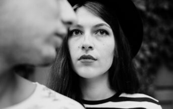 Black and white photo of a woman with long hair and a hat, looking intently at the camera. A person in the foreground, out of focus, partially obscures the left side of the image. The background is blurred.