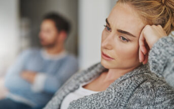 A woman with blonde hair, wearing a gray sweater, rests her head on her hand and looks pensive. A man in the background, who is out of focus, appears to be sitting with his arms crossed, wearing a blue and white striped shirt.