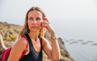 A woman with blonde hair wearing a black tank top and red backpack sits thoughtfully on a rocky cliff overlooking the ocean. She rests her chin on her hand, gazing into the distance. The ocean and a hazy sky are visible in the background.