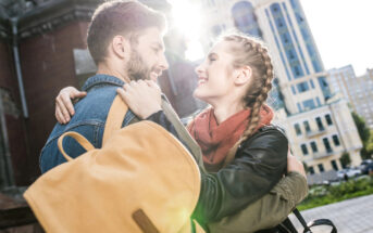 A smiling couple embraces outdoors with a cityscape in the background. The woman, with braided hair and wearing a scarf and leather jacket, looks at the man, who is wearing a denim jacket and yellow backpack. Sunlight filters through the buildings behind them.