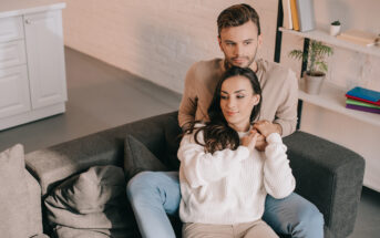 A man and woman sit on a dark gray couch in a cozy, modern living room. The woman is sitting in front of the man, both dressed in casual, light-colored clothing. The man gently embraces the woman from behind. A white bookshelf with books and a small plant is in the background.