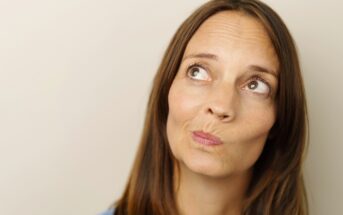 A woman with long brown hair is looking up and to the side with a thoughtful expression on her face. She is wearing a light blue top, and the background is a plain, light-colored wall.