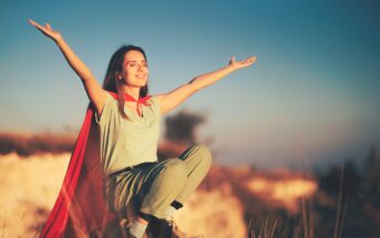 A woman wearing a red cape and green clothes stands on a rocky landscape with her arms outstretched, smiling and basking in the sunlight. The background is a blurred view of nature, with the warm glow of the setting sun illuminating the scene.