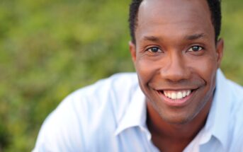 A man with short curly hair is smiling at the camera. He is wearing a light blue collared shirt. The background is blurred greenery.