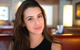 A woman with long, brown hair and a slight smile is sitting indoors. She is wearing a sleeveless black top. The background is softly blurred, showing a warm, well-lit interior with hanging lights and some blurred figures in the distance.