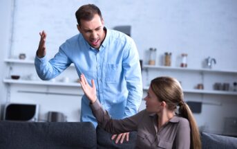 A man and a woman are having an intense argument in a living room. The man, standing and dressed in a blue shirt, is raising his hand and appears frustrated. The woman, sitting on a couch and wearing a gray shirt, is gesturing back at him, looking upset.