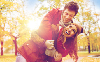 A young couple dressed in autumn clothing is playfully embracing in a park surrounded by colorful fall foliage. The man, in a red jacket, is leaning over the smiling woman, who is wearing a hat and scarf. They appear happy and joyful in the sunny, vibrant setting.