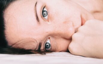 Close-up of a person's face lying down with their cheek resting on a surface. The individual's blue eyes are gazing intently towards the camera, and their hand is partially covering the lower half of their face.