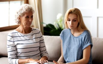 An older woman with curly gray hair and a younger woman with long blonde hair are sitting closely together on a beige sofa. Both appear to be deep in thought or concerned. The room is bright, with sunlight streaming in through a window in the background.