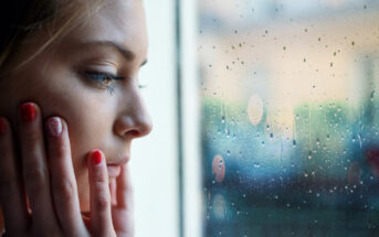 Close-up of a person with long hair looking out a window with raindrops on it. They are touching their face with both hands, and their expression appears contemplative. The background is blurred. Their nails are painted red, with a decorative design on one nail.
