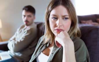 A woman with blonde hair sits on a couch, looking thoughtful and slightly distressed, with her hand resting on her chin. In the background, a man with a beard sits with his arms crossed, gazing towards her with a serious expression. The setting appears to be a living room.