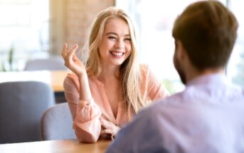 A woman with long blonde hair, wearing a peach blouse, smiles and gestures with her hand while talking to a man with short brown hair, sitting across from her at a wooden table in a brightly lit restaurant or cafe.