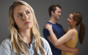 A woman with long blonde hair and a blue shirt looks upset in the foreground. In the background, a couple is embracing and smiling at each other. The woman's facial expression suggests feelings of sadness or jealousy.