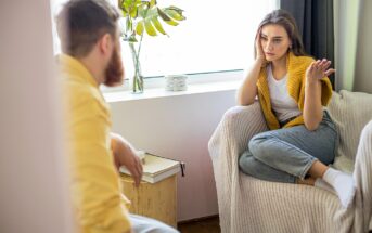 A woman sits on a chair with a frustrated expression and hand gesture while talking to a man seated nearby, who is facing her. The man has a beard and is wearing a yellow shirt. They are in a bright room with a window and plants in the background.