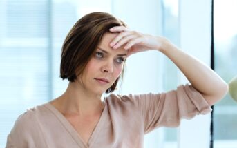 A woman with short brown hair, wearing a light peach blouse, stands by a window. She appears concerned or reflective, resting her forehead on her right hand while leaning slightly to the side. Natural light filters through the window behind her.