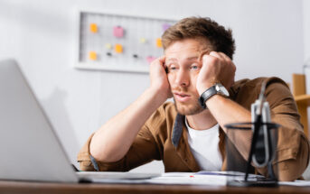A man with a frustrated expression sits at a desk, staring at a laptop screen. He rests his head in his hands, with elbows on the desk. In the background, there's a board with sticky notes attached. A pen holder and some papers are on the desk in front of him.