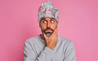 A man with a beard wearing a grey sweater and a tinfoil hat is looking to the side with a thoughtful expression, holding his chin. The background is pink.
