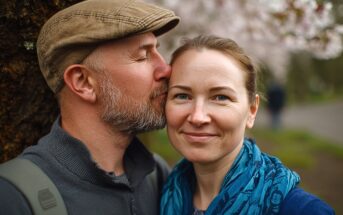 A man wearing a cap kisses a smiling woman on the cheek as they stand outdoors near a tree, with blurred greenery and blossoming trees in the background.