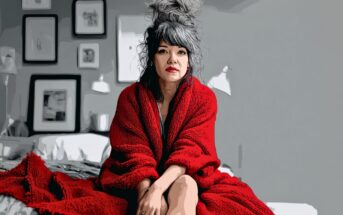 A woman with gray hair in a messy bun sits on a bed, wrapped in a bright red blanket. The room has gray walls with framed artwork. The overall image has a painted, stylized effect.