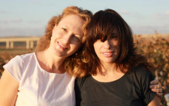 Two women stand outdoors, smiling at the camera. The woman on the left has light curly hair and is wearing a white top, while the woman on the right has dark hair with bangs and is wearing a black top. They have their arms around each other, with a scenic background behind them.