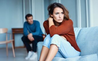 A woman sits on a couch looking upset with her arms around her knees, while a man sits in the background, both appearing distant and troubled in a modern living room.