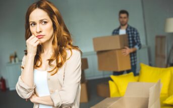 A woman with long brown hair looks thoughtful and concerned in the foreground. Behind her, a man carries moving boxes in a room with yellow cushions on the floor. The scene suggests a moving or packing scenario.