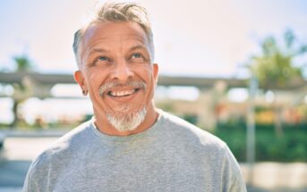 A middle-aged man with gray hair and a beard smiles while looking up. He is wearing a gray T-shirt and standing outdoors, with a blurred background of greenery and structures on a sunny day.