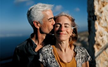 A man with gray hair gently kisses a smiling woman on the forehead. They stand outdoors by a stone wall, bathed in sunlight, with a blue sky and ocean in the background.