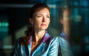 A woman in a blue patterned blazer gazes thoughtfully out a window, with sunlight illuminating her face and her reflection visible in the glass beside her.