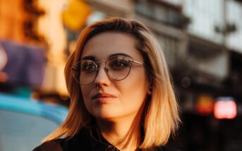 A person with short blonde hair, wearing round glasses and a nose ring, looks thoughtfully into the distance. The background shows an urban street scene bathed in warm sunlight.