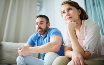 A woman and a man sit on a couch indoors, both looking serious and thoughtful. The woman is resting her chin on her hand, while the man sits with his hands clasped, gazing forward.