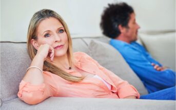 A woman in a peach shirt sits on a couch, looking thoughtful and slightly concerned, with her hand resting on her chin. In the background, a man in a blue shirt sits turned away, appearing distant.