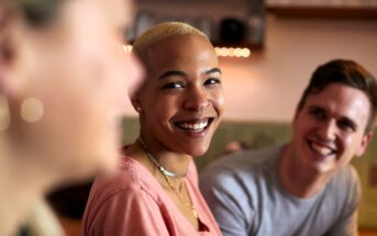 Three young adults are sitting together indoors, smiling and laughing. The focus is on a person with short blond hair and a nose ring, wearing a pink shirt, while the others are slightly out of focus in the foreground and background.