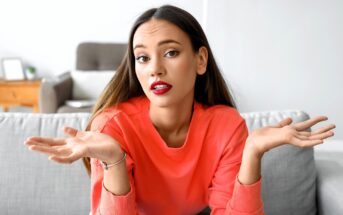A young woman with long brown hair and red lipstick sits on a gray couch, wearing a coral sweatshirt, and raises both hands with an unsure or questioning expression. A blurred living room is in the background.