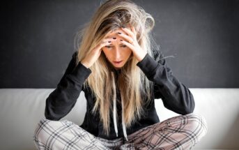 A young woman with long blonde hair sits cross-legged on a couch, wearing plaid pajama pants and a black hoodie, holding her head in her hands and looking down, appearing stressed or worried.