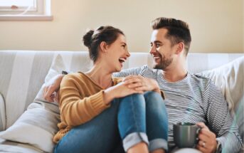 A smiling couple sits close together on a couch, laughing and looking at each other. One person wears a mustard sweater and jeans, while the other wears a striped shirt and holds a mug. The atmosphere appears warm and relaxed.