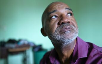 A middle-aged man with a graying beard and mustache looks thoughtfully upward. He is wearing a maroon shirt and is indoors, with a soft green background and some blurred furniture behind him.