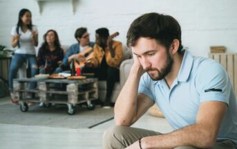 A man sits apart, looking sad and thoughtful, while a group of four people in the background sing, play guitar, and enjoy each other's company in a cozy living room.