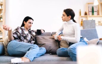 Two women sit on a couch having a serious conversation, each holding a mug. One gestures with her hand while speaking, and the other listens attentively. Both are dressed casually in jeans and tops.