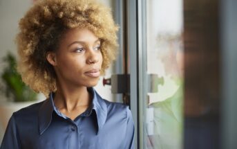 A woman with curly blonde hair and a nose ring gazes thoughtfully out of a window. She is wearing a shiny blue button-up shirt, and soft natural light illuminates her face. A blurred plant is visible in the background.