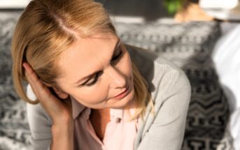 A middle-aged woman with blonde hair looks down thoughtfully while touching her hair, sitting indoors on a patterned blanket in soft natural light.