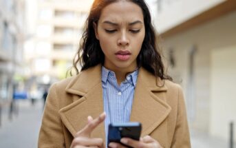 A woman with long dark hair wearing a tan coat and striped shirt looks at her smartphone with a concerned expression while standing outdoors on a city street.