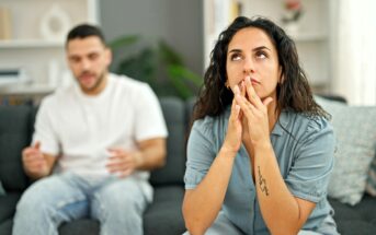 A woman sits on a couch looking frustrated and thoughtful, with her hands near her face, while a man in the background appears to be talking to her, looking concerned.
