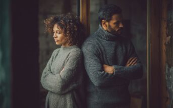 A woman and a man stand back to back with arms crossed, both looking away with serious expressions. They are indoors, wearing warm sweaters, and appear to be upset or in disagreement.